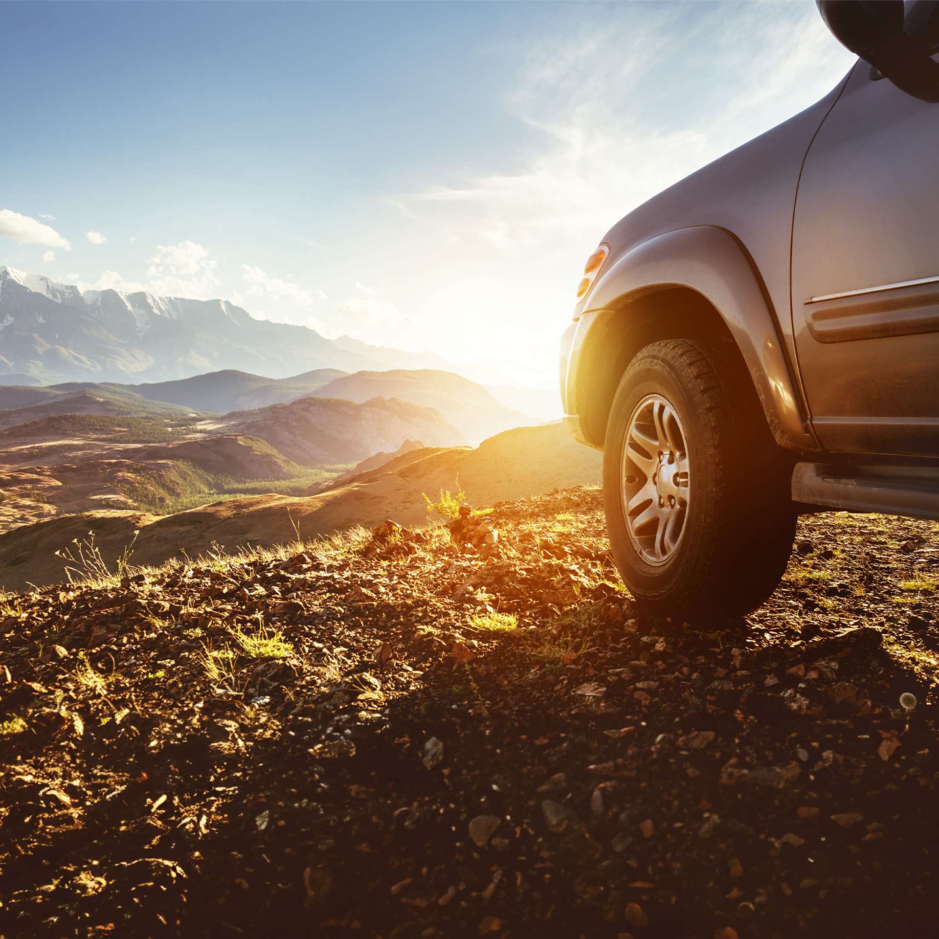 Ein SUV steht auf einem felsigen Weg mit Blick auf ein weites Bergpanorama und eine untergehende Sonne.