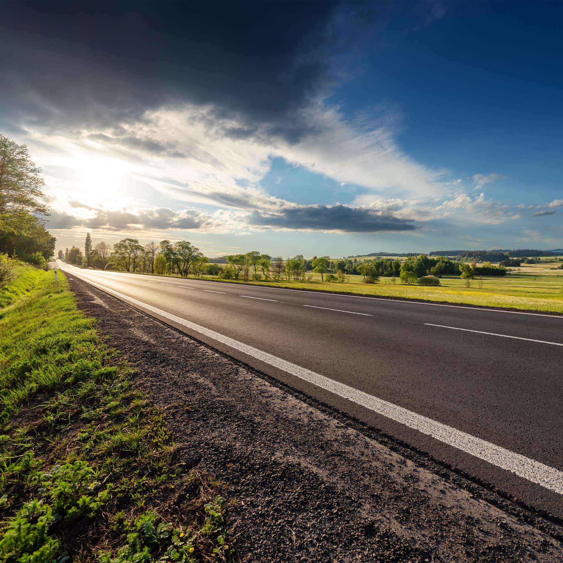  Eine leere Landstraße, die durch eine Landschaft mit dramatischem Himmel und untergehender Sonne führt.