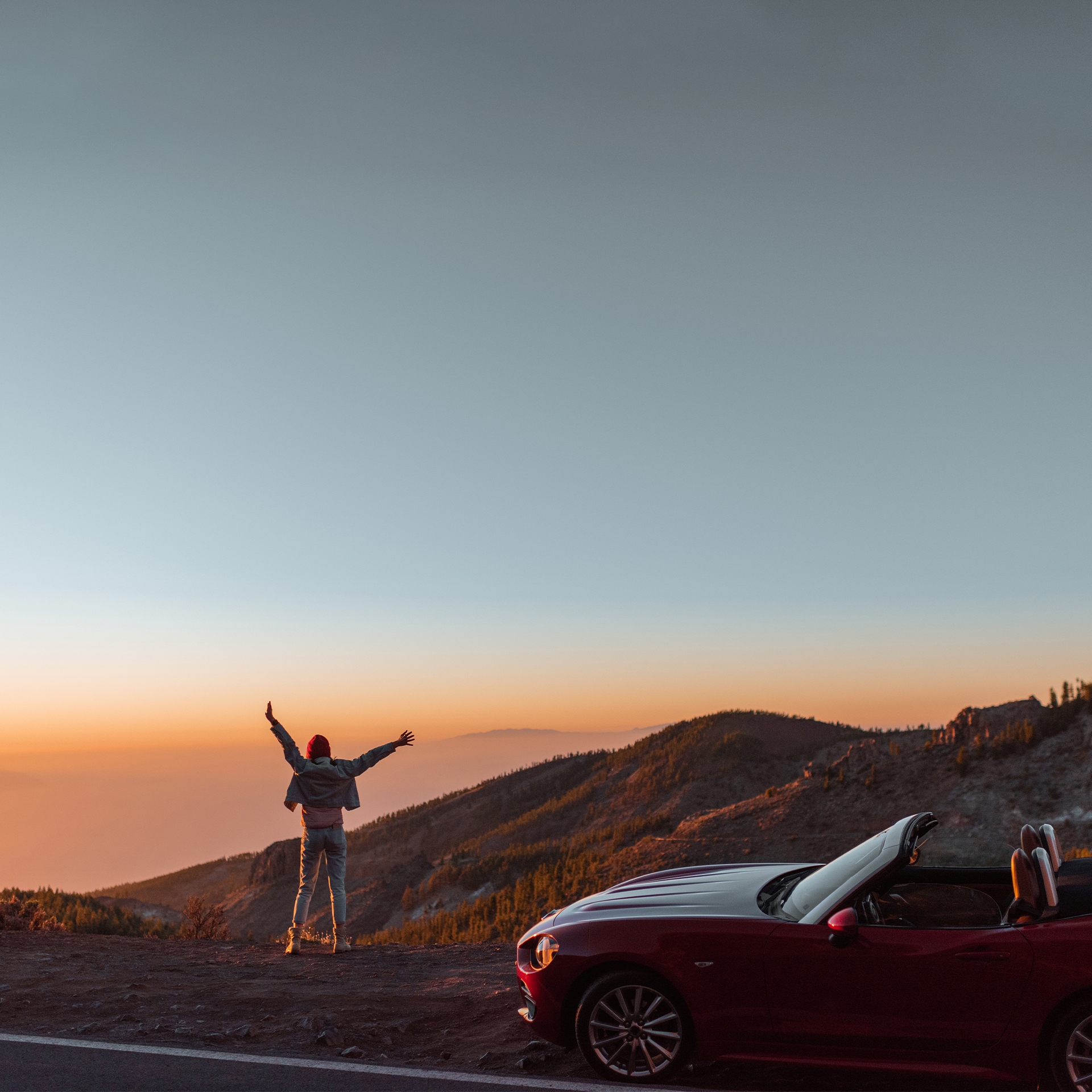 Ein rotes Cabrio parkt an einem Bergpass, während eine Person mit ausgestreckten Armen den atemberaubenden Sonnenuntergang und die umliegende Landschaft genießt.
