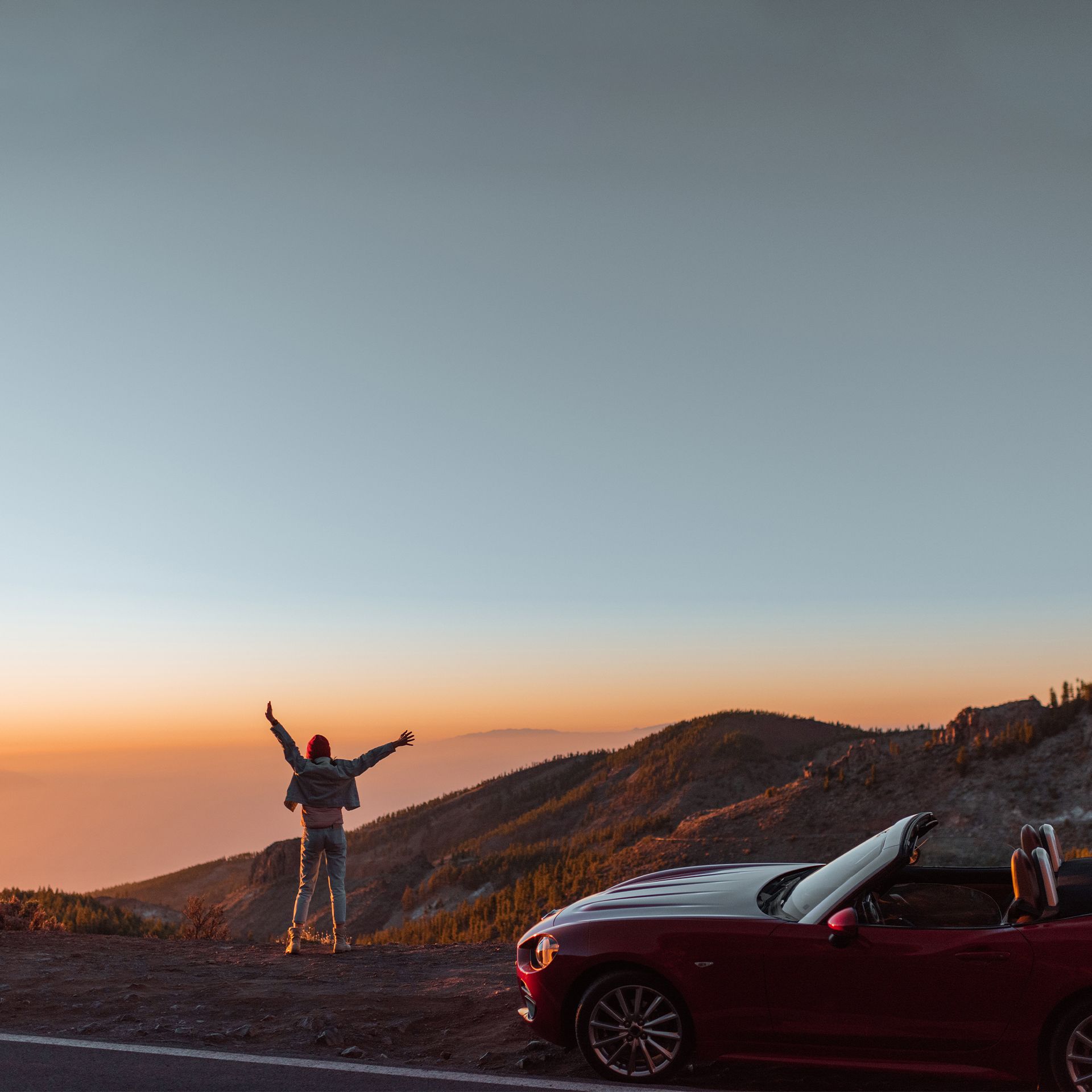 Ein rotes Cabrio parkt an einem Bergpass, während eine Person mit ausgestreckten Armen den atemberaubenden Sonnenuntergang und die umliegende Landschaft genießt.