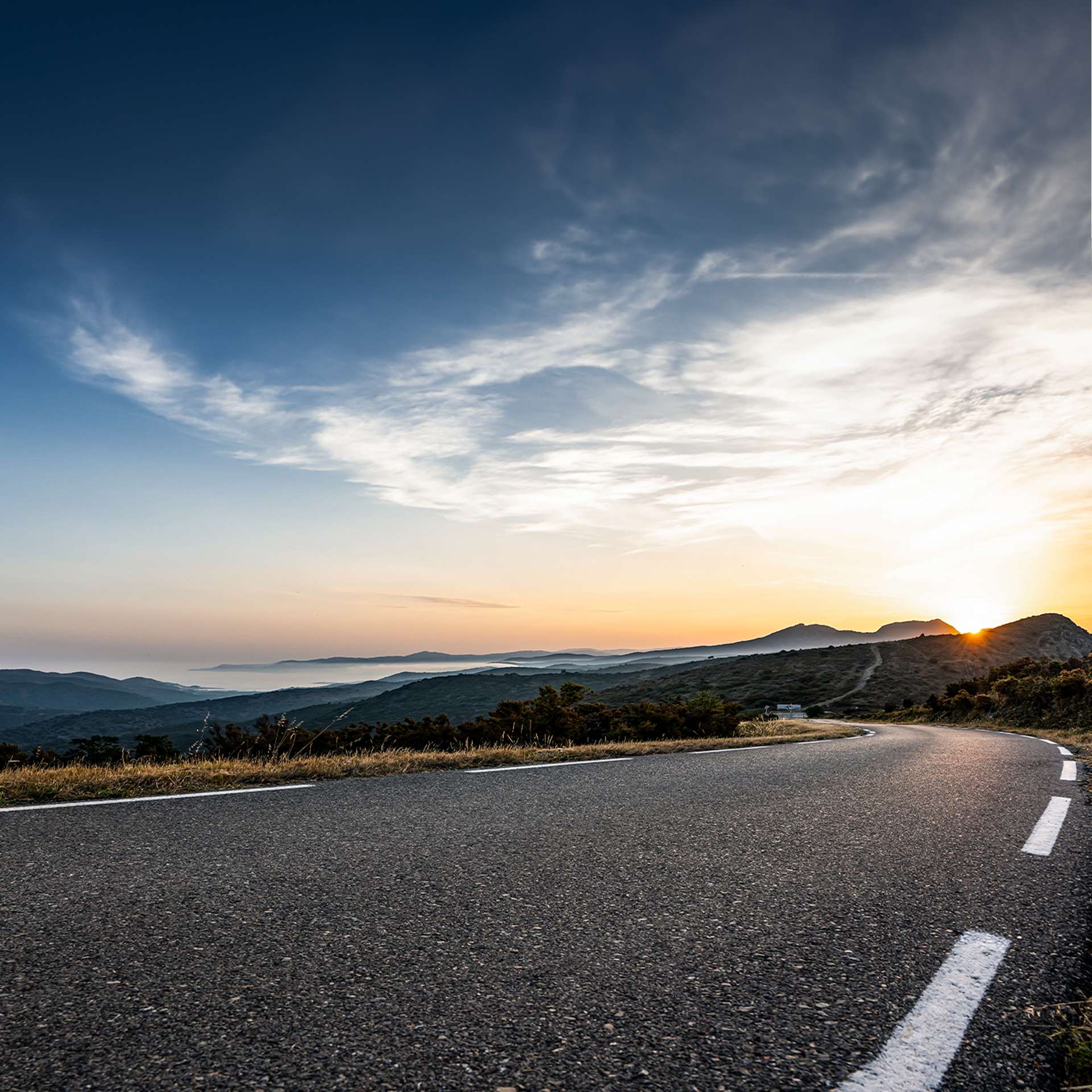 ine kurvige Straße mit Blick auf die Berge und das Meer bei Sonnenuntergang.