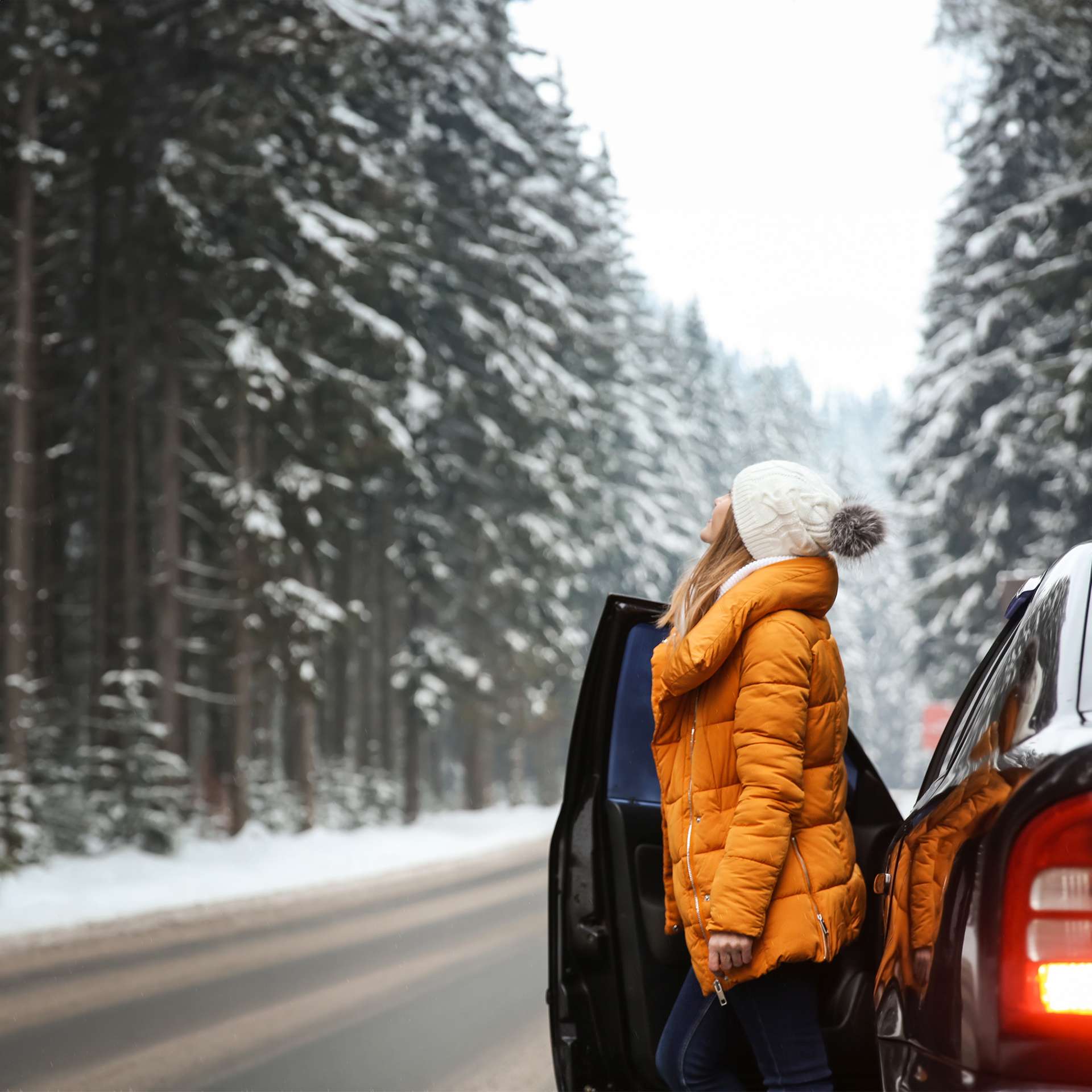 Eine Frau in gelber Winterjacke lehnt an ihrem Auto und blickt in die verschneite Waldlandschaft.
