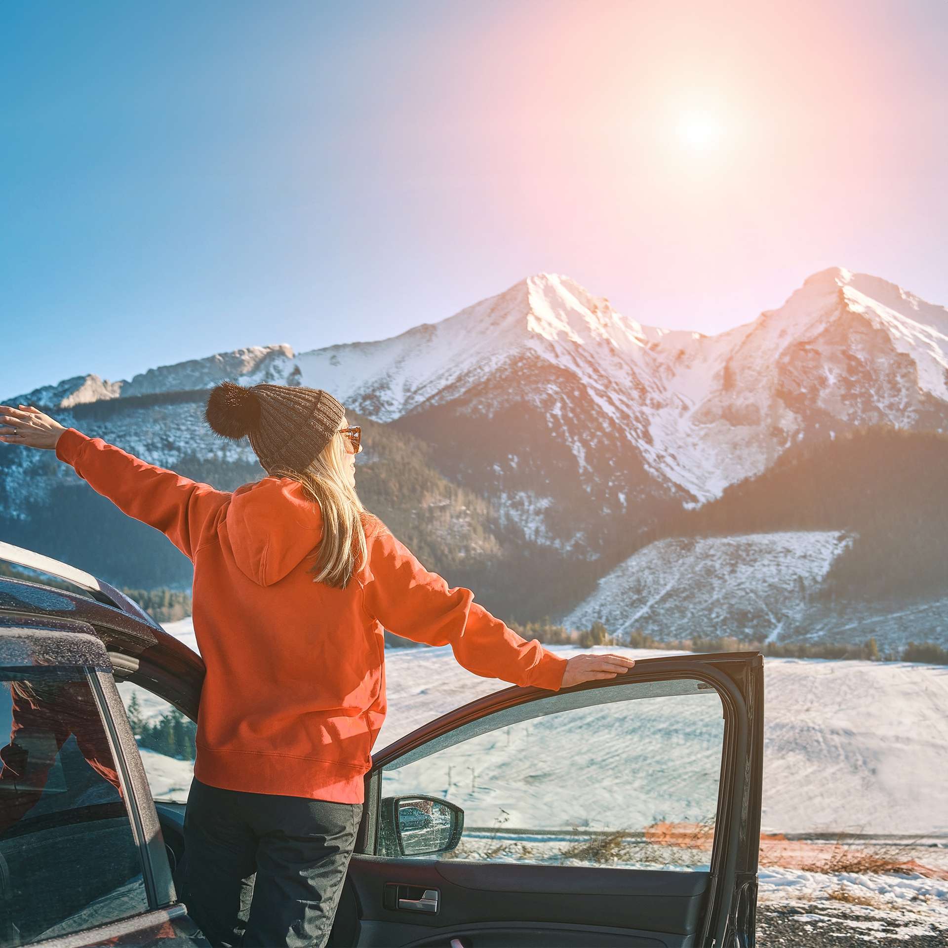 Eine Frau steht mit geöffneten Autotüren vor einer schneebedeckten Berglandschaft und genießt die Sonne.