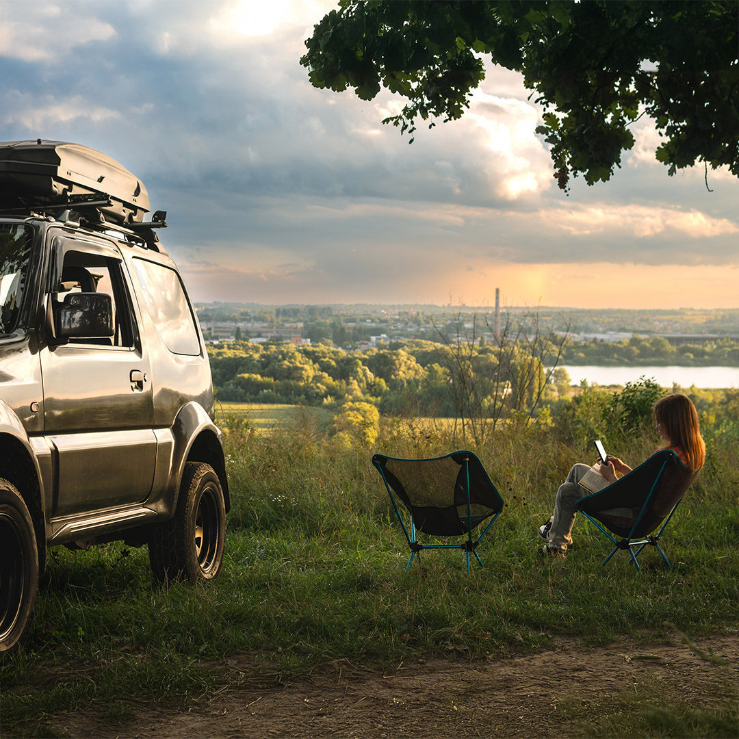 Two folding light weight, portable chairs and woman reading the book. Nature, on the top, river, sunset. Tourism and active lifestyle. Equipment for tourism. Off road vehicle with roof rack
